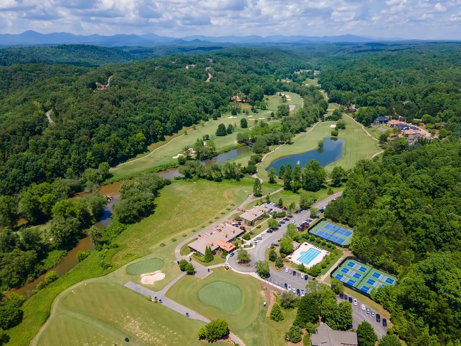 Wide aerial drone view of the Achasta community core in Dahlonega Georgia — clubhouse, tennis and pickleball courts, swimming pool, golf course, Chestatee River, and Blue Ridge foothills