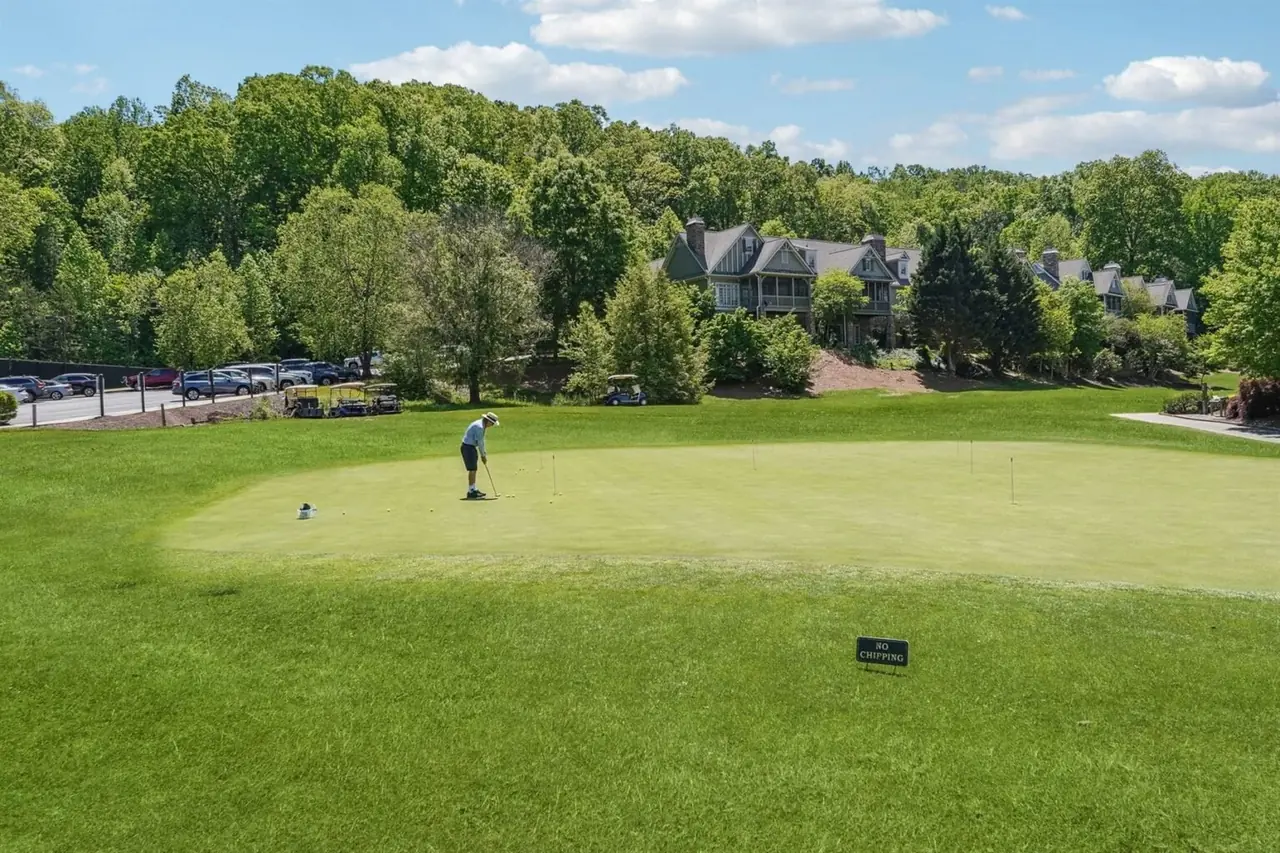 A single golfer practices on the Achasta putting green with a large gabled stone and shake estate home in the background