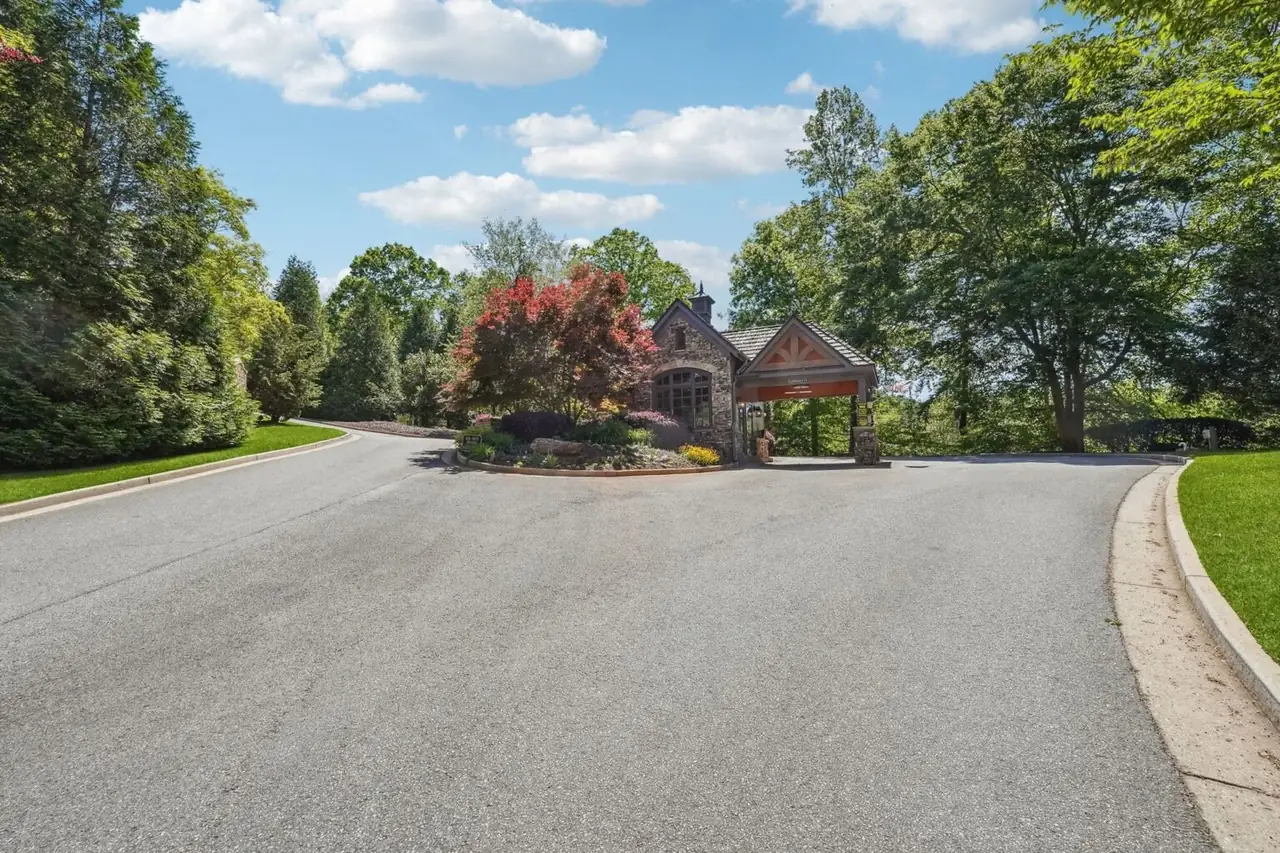 Achasta primary gate — stone gatehouse with timber porte-cochère for the gate keeper, ornamental landscaping with red-leaf shrubs in the median, and a wide paved entry sweep